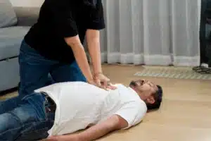 Person performing chest compressions on a man lying on the floor during a medical emergency.