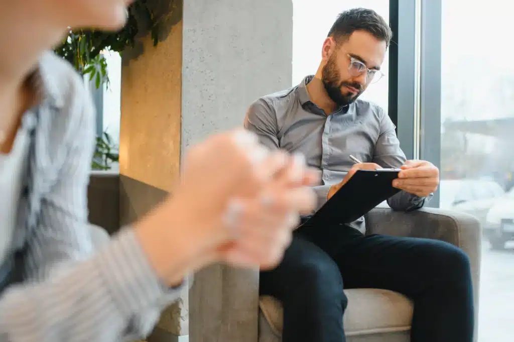 Therapist taking notes on a clipboard during a counseling session with a client in an office setting.