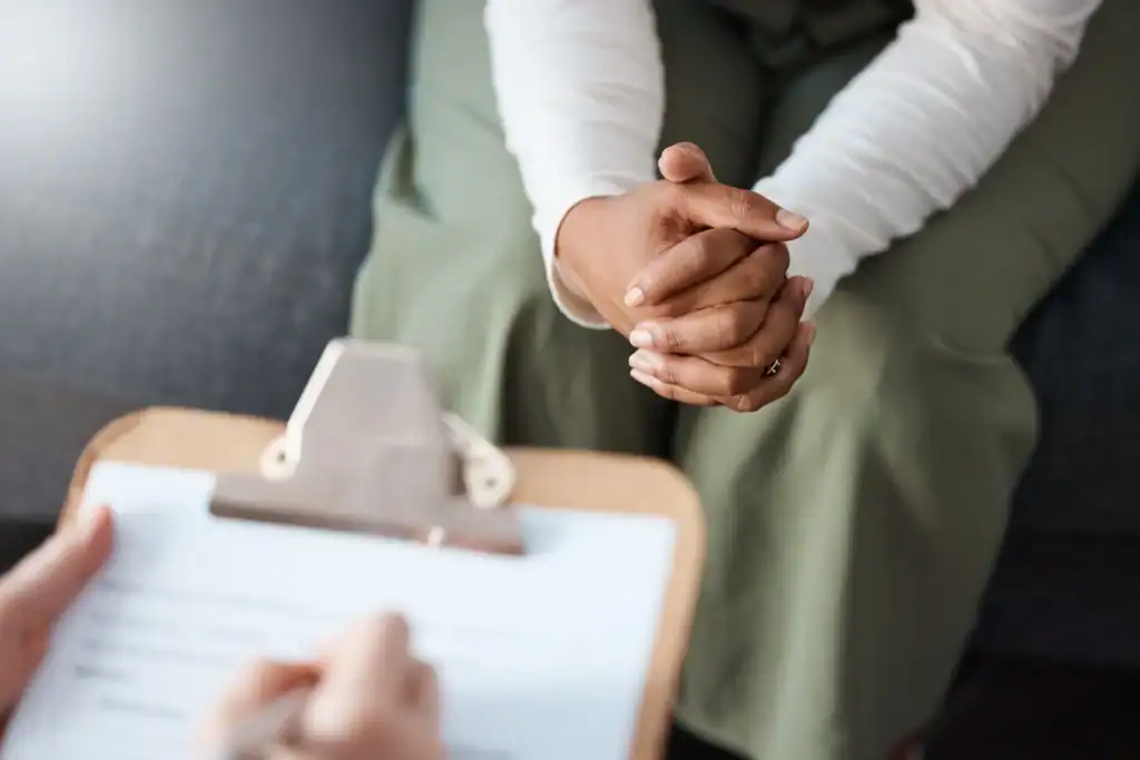 Clinician holding a clipboard during a drug and alcohol assessment while a person sits with hands clasped across from them.