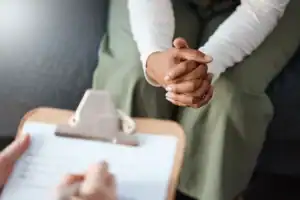 Clinician holding a clipboard during a drug and alcohol assessment while a person sits with hands clasped across from them.